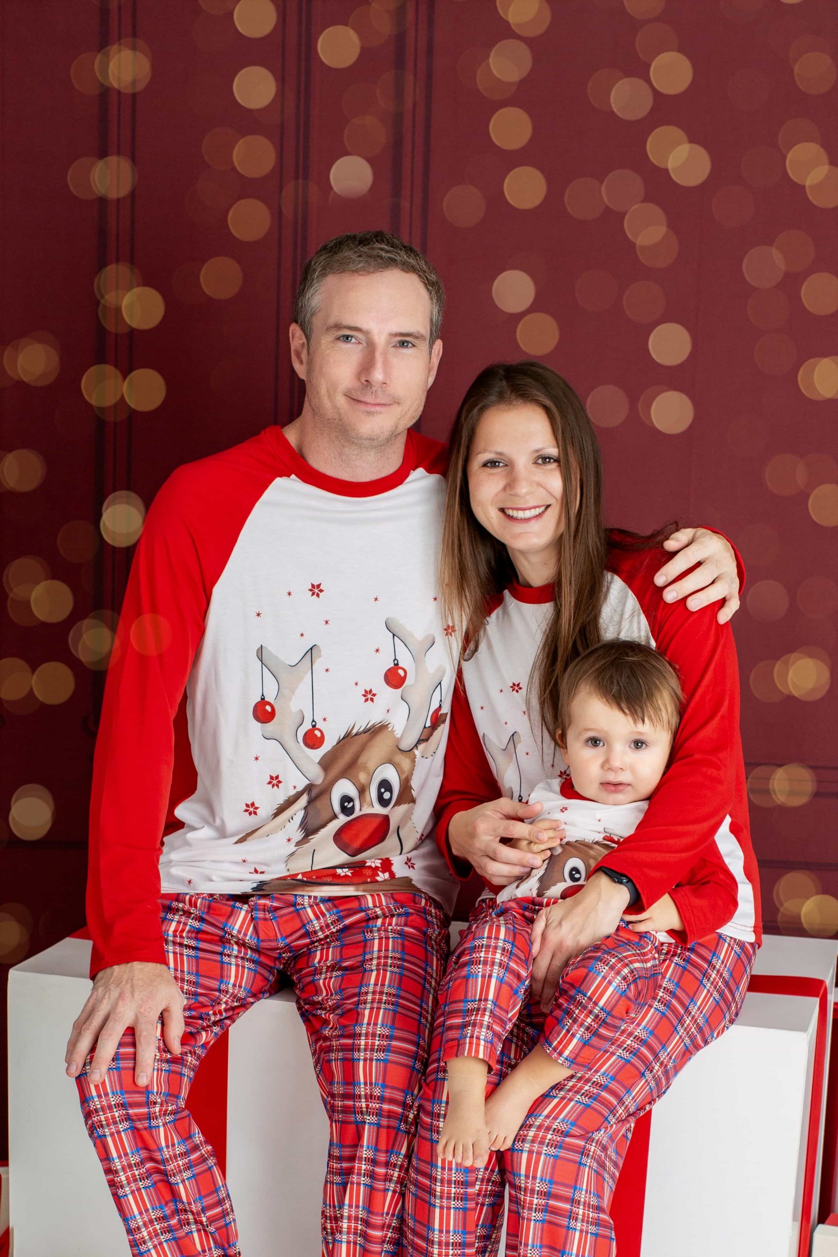A Family (2 adults and 1 baby) photographed on a Christmas backdrop, sitting on giant Christmas presents, smiling and looking straight to the camera