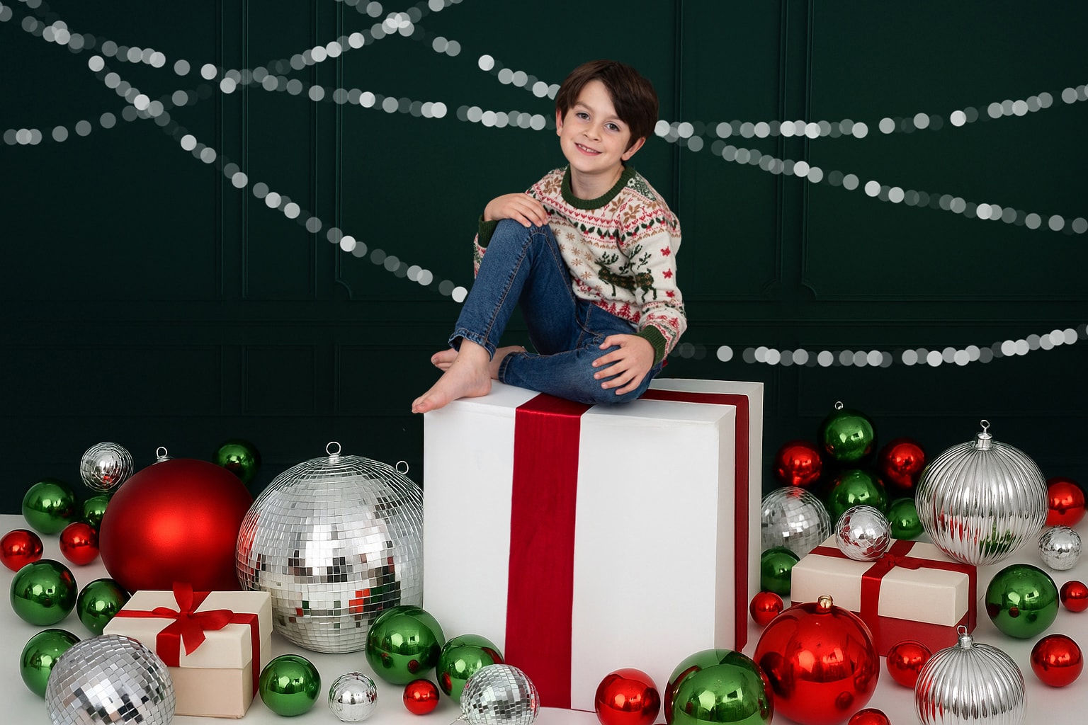 A young boy sits on a giant wrapped present surrounded by baubles and disco balls, with a green background and silver sparkles. Photo by Sarah Oliver Photographer at So Studios, Epping, Essex.